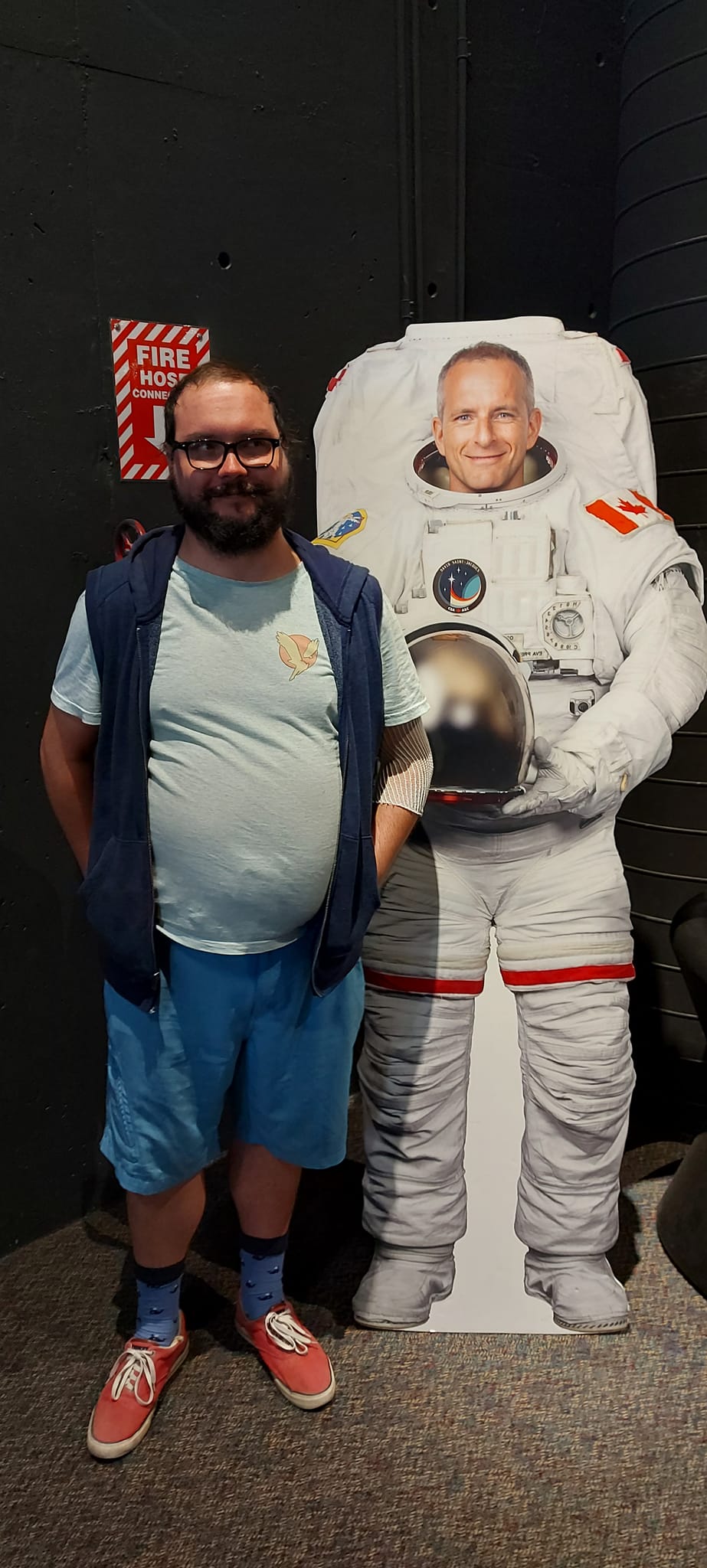 A man with a ball cap and bushy facial hair is standing next to a cardboard cut out of Christ Hadfield in a space suit